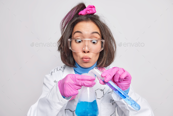 Indoor shot of funny surprised female chemist conducts chemical experiment mixes two reagents gets b