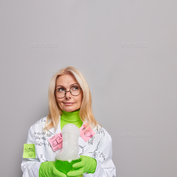 Vertical shot of pensive middle aged woman scientist holds glass flask with green liquid conducts ph