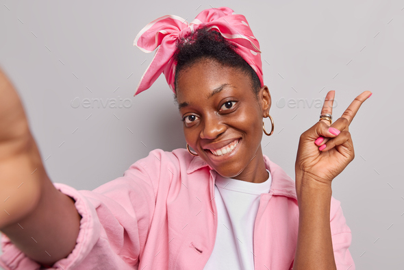 Good looking dark skinned woman makes peace gesture takes selfie portrait smiles gently wears kerchi