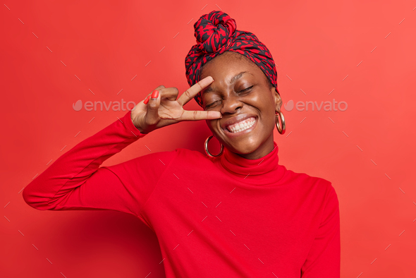 Optimistic dark skinned Afro American woman shows v sign near face peace gesture keeps eyes closed s