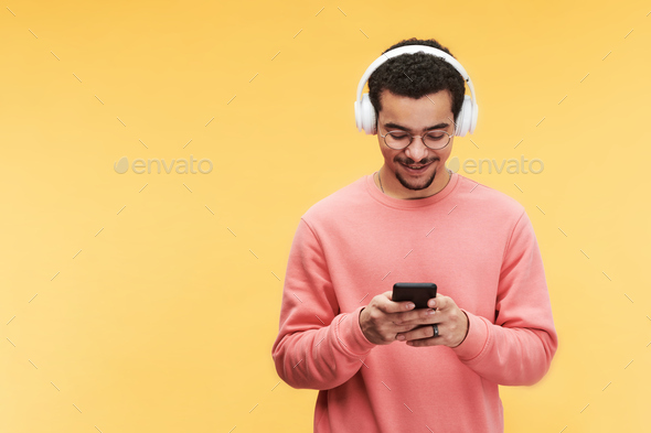 Happy guy in headphones, eyeglasses and pink sweatshirt using smartphone