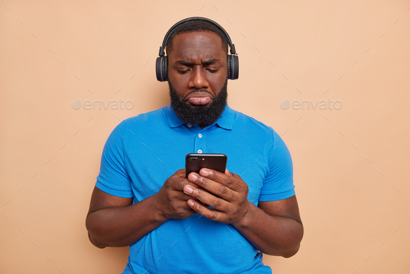 Displeased dark skinned man with thick beard types text messages via smartphone unhappy to read bad