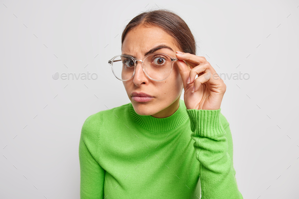 Portrait of attentive young European woman keeps hand on rim of spectacles has focused gaze at camer