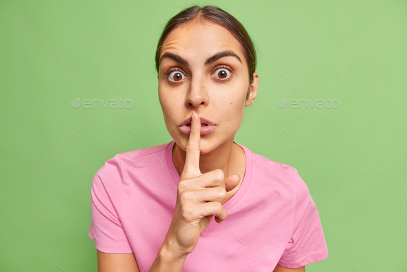 Shush its secret. Surprised brunette woman tells rumors makes silence gesture holds index finger on