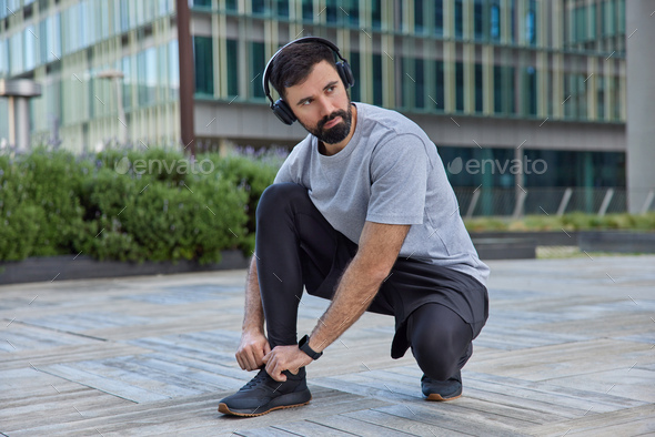 Active healthy man prepares for jogging laces shoes looks away with thoughtful expression listens mu