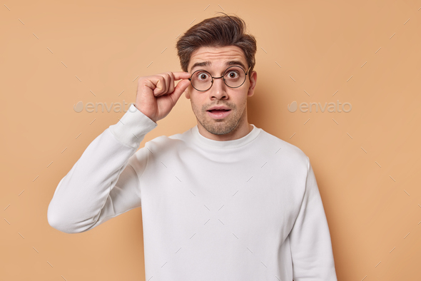 Studio shot of amazed brunet man keeps hand on rim of spectacles looks at something unbelievable can