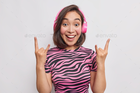 Positive Asian woman makes rock gesture enjoys listening music in wireless headphones shows heavy me