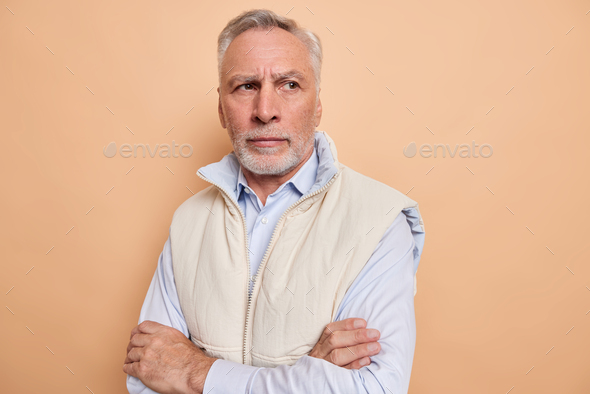 Indoor shot of thoughtful elderly man keeps arms folded thinks of personal problems lost in thoughts
