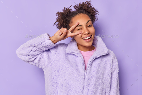 Joyful young woman with hair buns winks eyes makes peace sign over eye smiles happily wears fur coat
