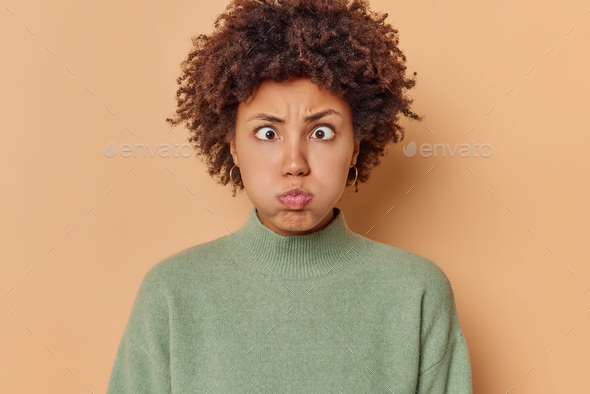Photo of funny curly haired young woman pouts lips blows cheeks makes hilarious grimace holds breath