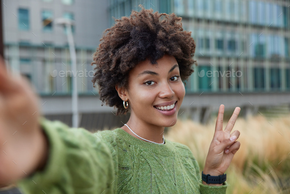 Happy young woman enjoys spare time makes peace sign stretches arm makes selfie smiles gladfully wea