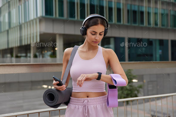 Outdoor shot of sporty fit woman poses with sport equipment checks time on smartwatch uses smartphon