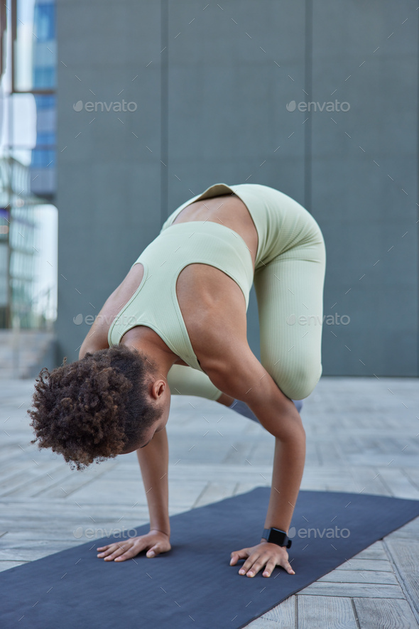 Active slim woman with curly combed hair dressed in cropped top and leggings leans arms on fitness m