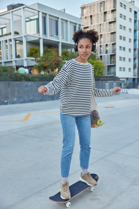Full length shot of active teenage girl dressed in casual clothes rides skateboard listens music via