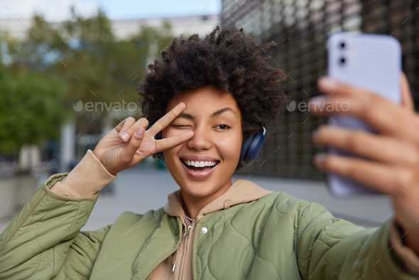 Beautiful smiling woman makes peace gesture over eye has carefree expression takes selfie via smartp