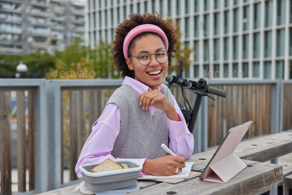 Smiling content woman browses tablet and writes down notes sin notebook makes content plan for blog