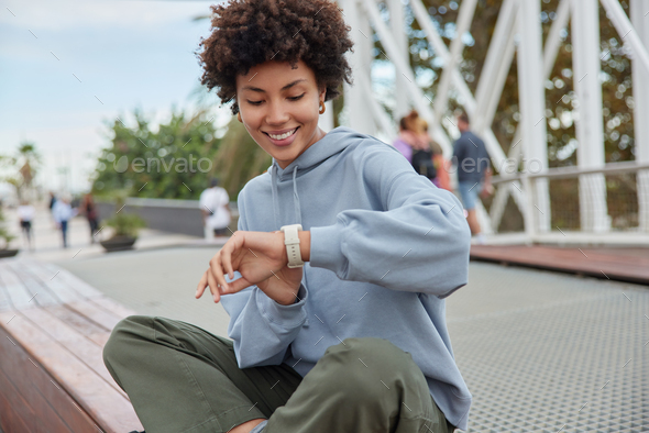 Outdoor shot of happy curly haired woman checks covered distance on smartwatch sits crossed legs aga