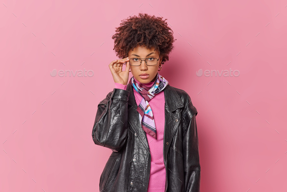 Portrait of serious self assured young woman keeps hand on rim of spectacles looks at camera feels c