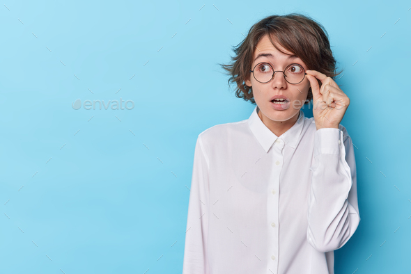 Horizontal shot of stunned woman stands speechless holds breath keeps hand on rim of spectacles wear