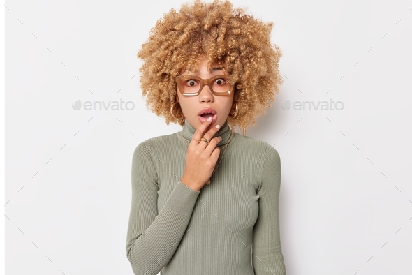 Horizontal shot of shocked curly haired European woman keeps mouth opened holds breath cannot believ