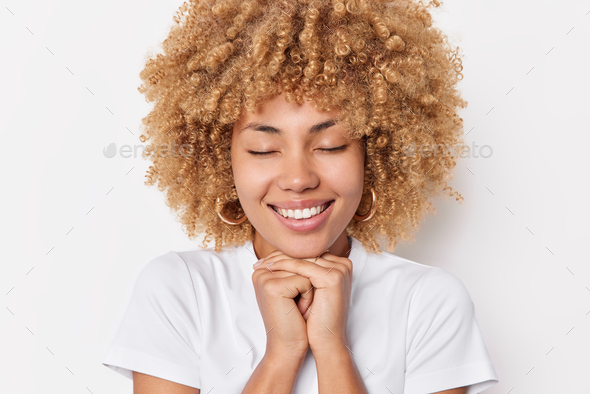 Dreamy Curly Haired Young Woman Keeps Hands Under Chin Smiles Pleasantly Recalls Something 