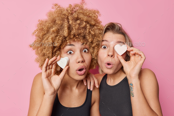 Two surprised women hold heart shaped soaps take care of skin and complexion look with widely opened