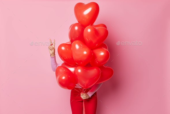 Hide and seek concept. Unknown woman makes peace gesture hides behind red inflated heart balloons ma