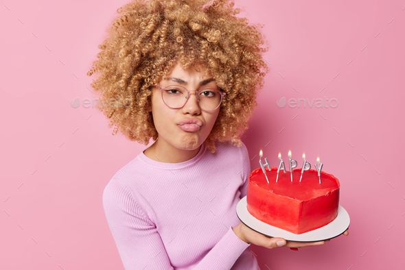 Serious woman makes unhappy grimace purses lips holds tasty heart shaped cake prepares for Valentine