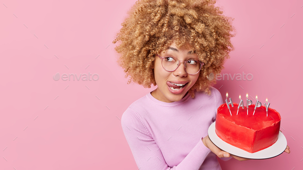 Horizontal shot of pretty curly haired woman sticks out tongue holds delicious red heart shaped cake