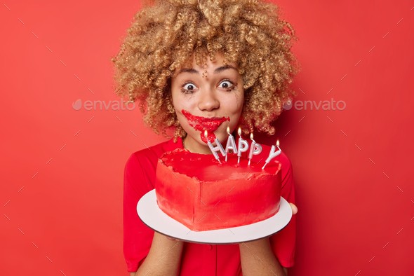 Surprised curly haired woman make mess eating big fancy heart shaped cake has mouth smeared with cre