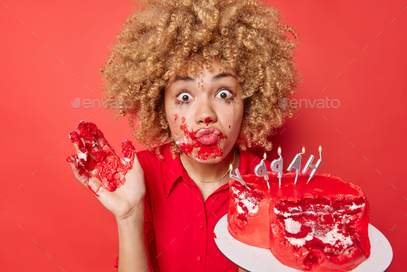 Shocked curly haired woman smeared with cream after eating cake looks surprised at camera keeps lips