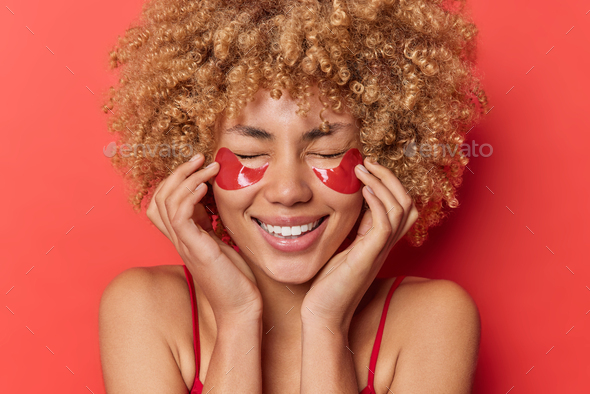 Optimistic cheerful curly haired woman applies collagen patches under eyes to reduce dark circles an