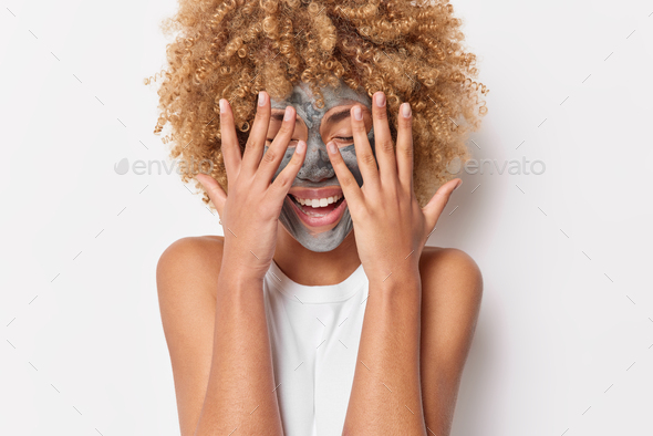 Positive young woman with curly bushy hair keeps hands over face tries to hide face appies clay mask