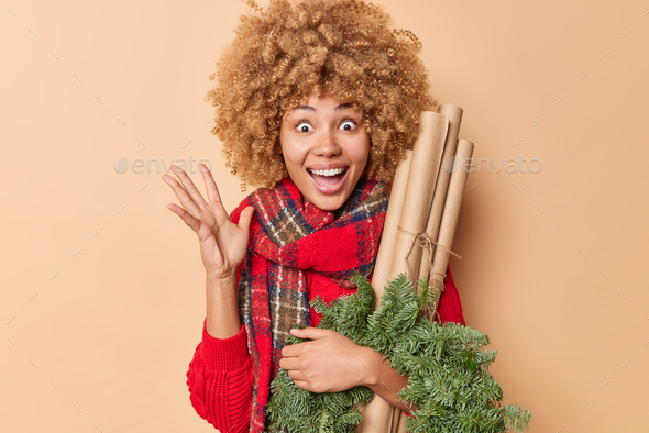 Overjoyed surprised woman reacts on nice news keeps hand raised carries green spruce wreath and roll