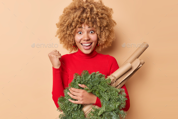 Joyful curly haired woman clenches fist and celebrates good news holds green spruce wreath and rolle