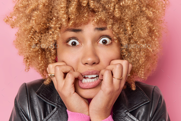 Headshot of worried curly haired woman bites finger nails looks nervously at camera afraids of somet