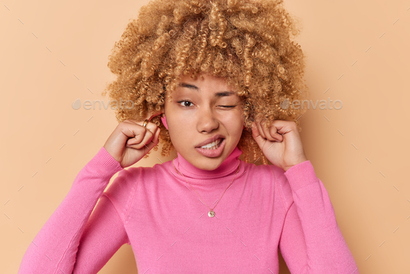 Too loud. Displeased curly haired young European woman doesnt want to hear something plugs ears igno