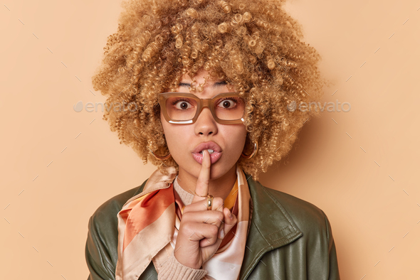 Surprised woman makes taboo gesture silence sign shushes indoor looks mysterious expression dressed