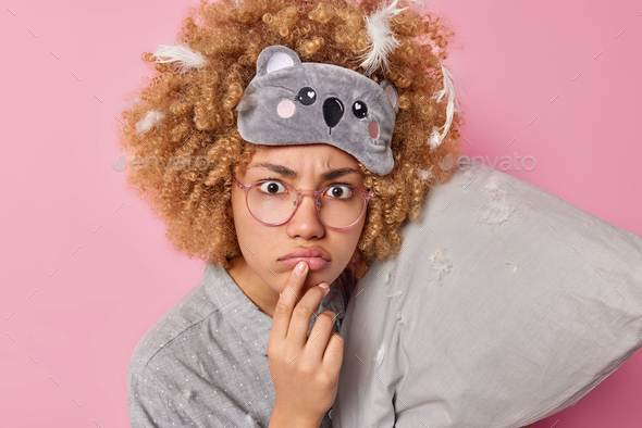 Photo of discontent curly haired young woman purses lips prepares for sleep wears blindfold on foreh