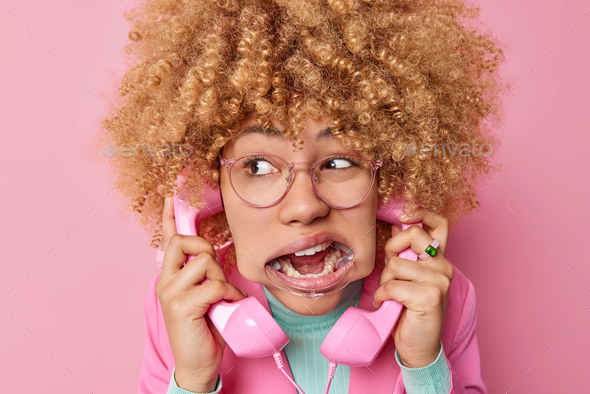 Close up shot of curly haired young woman talks via pink retro handset phone has widely opened mouth