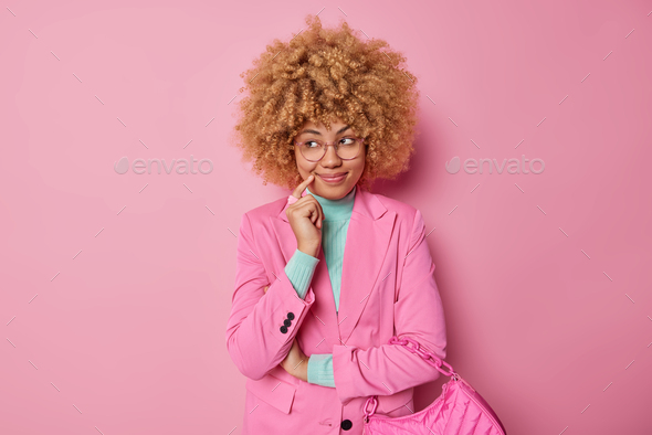 Pretty young woman with curly bushy hair looks away with dreamy satisfied expression makes plans and