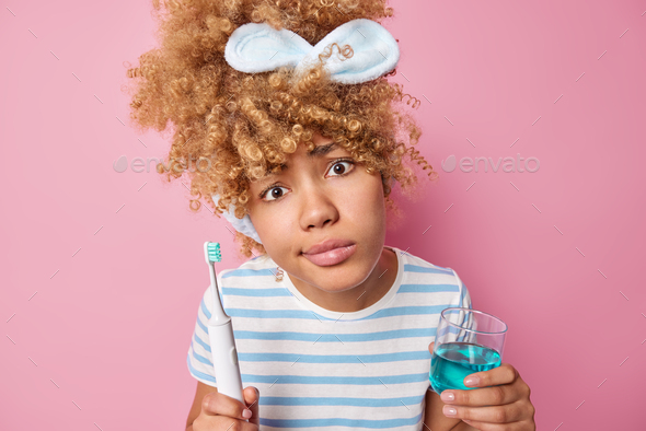 Indignant curly haired woman holds glass of mouthwash and toothbrush for fresh breath wears headband