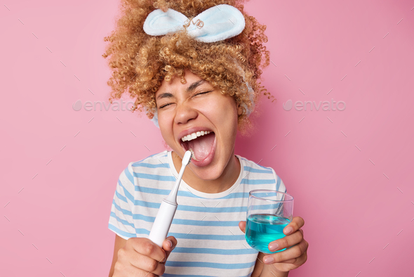 Optimistic curly haired girl keeps mouth widely opened brushes teeth ...