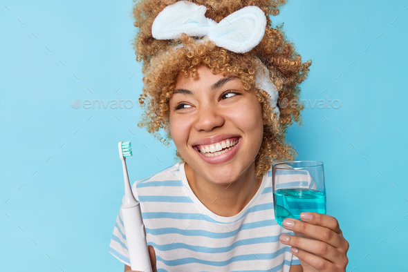 Positive young woman holds glass of mouthwash and electric toothbrush for fresh breath looks gladful