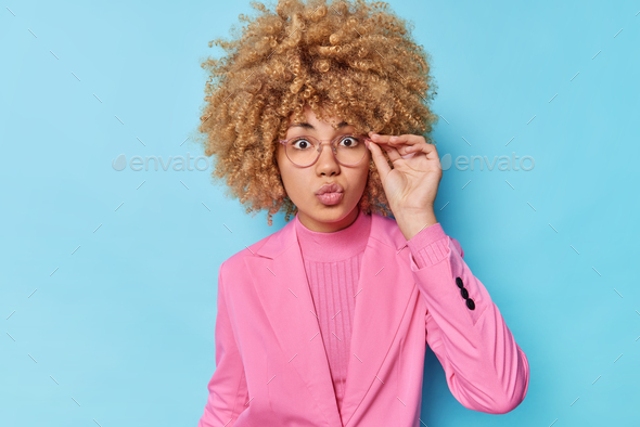 Serious wondered woman with curly bushy hair keeps lips folded hand on rim of spectacles dressed in