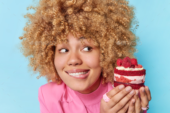 Curly haired beautiful woman bites lips looks at appetizing piece of cake with raspberries has sweet
