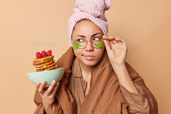 Serious young woman looks attentively aside keeps hand on rim of spectacles poses with delicious pan