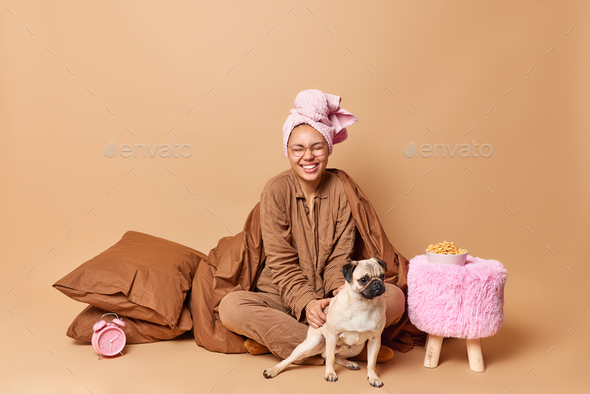 Positive young woman plays with pug dog after awakening wears nightwear and bath towel wrapped on he