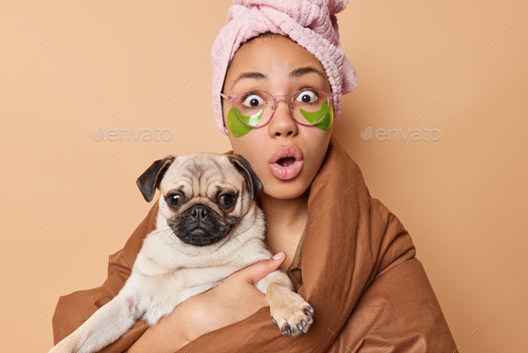 Indoor shot of shocked young woman stares bugged eyes keeps mouth opened poses with favorite pug dog