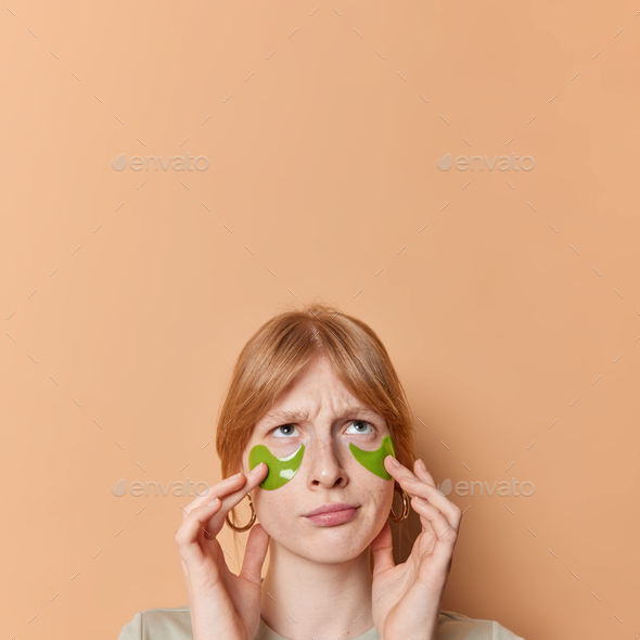 Vertical shot of frustrated ginger girl applies beauty patches under eyes to remove wrinkles frowns